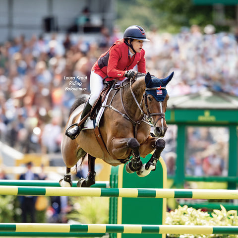 Laura Kraut Riding Baloutinue. Rider jumping a brown horse over a green and yellow jump during a competition with crowd in the background.
