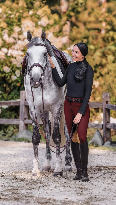 Rider in black turtleneck and maroon breeches standing next to a gray horse in fall setting