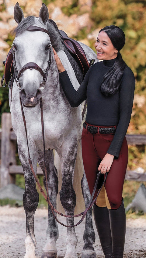 Rider in black turtleneck and maroon breeches standing next to a gray horse in fall setting