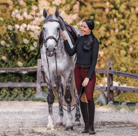 Female rider in black turtleneck and maroon breeches standing next to a gray and white horse in front of floral background with a fence.