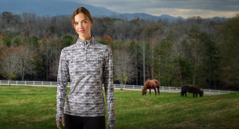Woman standing in a field wearing Ariat print long sleeve shirt with horses and mountains in the background