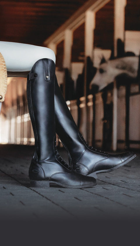 Riders legs wearing black tall boots seated in front of horse stalls