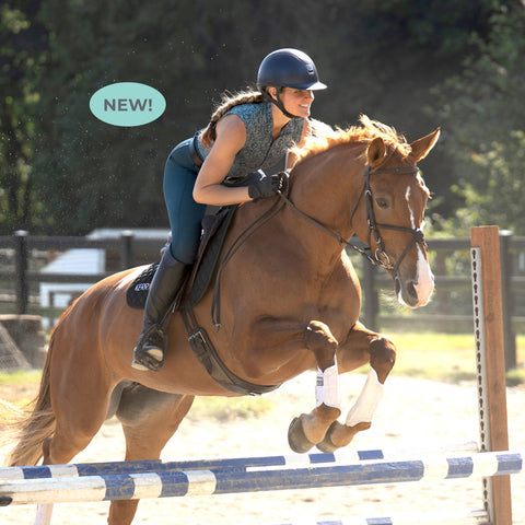 A woman is riding a horse over a jump while wearing a teal-hued quarter zip sleeveless top with a paisley pattern and matching teal breeches.