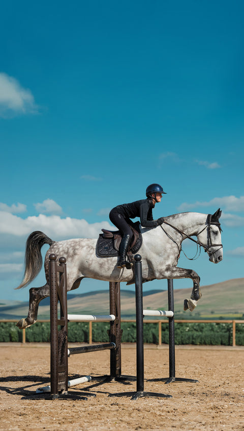 Person riding a gray horse over an oxer jump in an outdoor arena