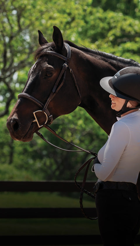 Woman, dressed to ride, smiling at dark bay horse with bridle on.