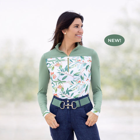 Smiling woman wearing a sage green long-sleeve equestrian top with a floral print front and quarter-zip collar, paired with dark riding breeches.